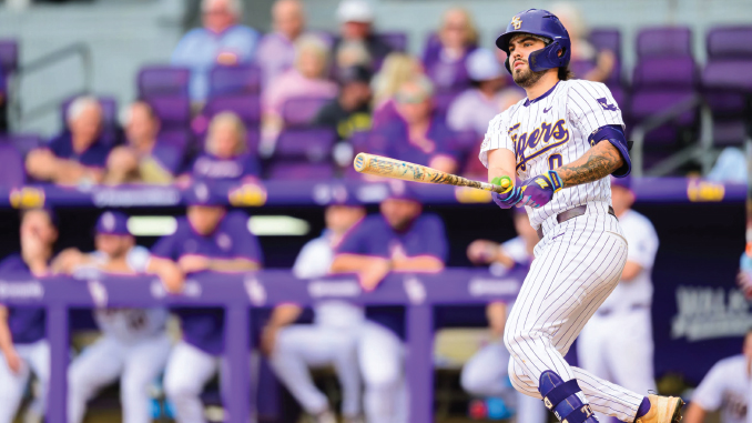 Baseball player in a white pinstripe uniform swings a bat at the plate, helmet on, during a game with a purple-dyed dugout in the background.