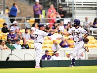 Two baseball players in white pinstriped uniforms high-five on the field as fans sit in yellow seats behind them.
