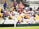 Two baseball players in white pinstriped uniforms high-five on the field as fans sit in yellow seats behind them.