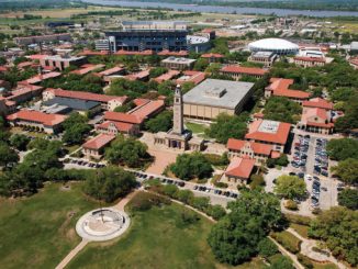Aerial view of a university campus with red-roofed buildings, a tall clock tower near a central plaza, a circular monument in a green park, and a large stadium in the distance.