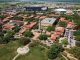 Aerial view of a university campus with red-roofed buildings, a tall clock tower near a central plaza, a circular monument in a green park, and a large stadium in the distance.