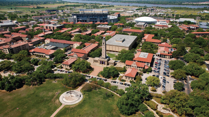 Aerial view of a university campus with red-roofed buildings, a tall clock tower near a central plaza, a circular monument in a green park, and a large stadium in the distance.