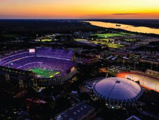 Aerial view of LSU Tiger Stadium at dusk, glowing purple and surrounded by a circular domed arena, with a river in the distance.