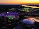 Aerial view of LSU Tiger Stadium at dusk, glowing purple and surrounded by a circular domed arena, with a river in the distance.