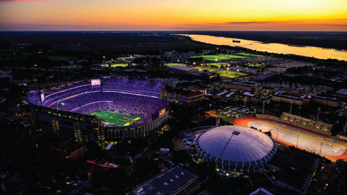 Aerial view of LSU Tiger Stadium at dusk, glowing purple and surrounded by a circular domed arena, with a river in the distance.