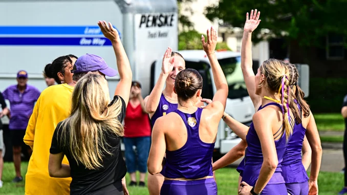 Female athletes in purple uniforms high-fiving in a circle on a grassy field, with a Penske truck in the background.