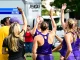Female athletes in purple uniforms high-fiving in a circle on a grassy field, with a Penske truck in the background.