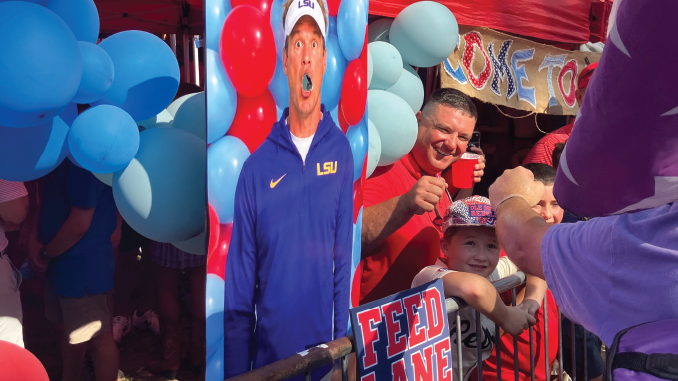 LSUFBall091926_GrovePreGame2_GGuilbeau | TigerRag.com Life-size cardboard cutout of a man in an LSU blue jacket among blue and red balloons at a festival booth, with cheering spectators nearby.