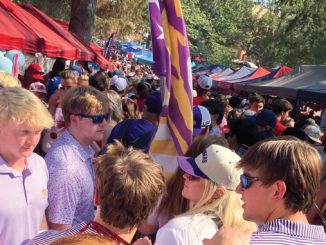 Busy outdoor market or festival scene with red and blue tents, balloons, and a crowd of people wearing sunglasses and hats.