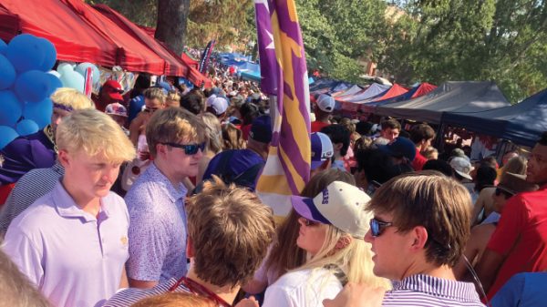Busy outdoor market or festival scene with red and blue tents, balloons, and a crowd of people wearing sunglasses and hats.