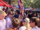 Busy outdoor market or festival scene with red and blue tents, balloons, and a crowd of people wearing sunglasses and hats.