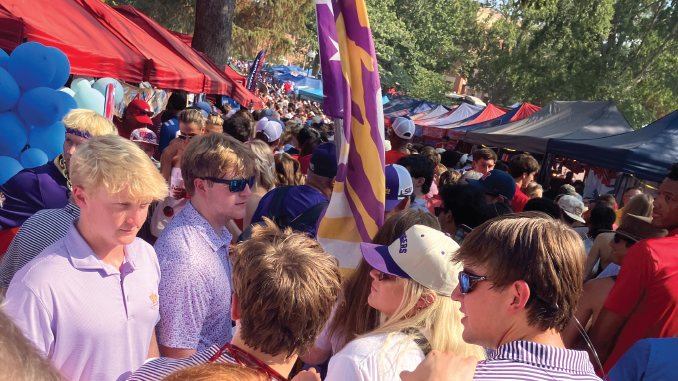 Busy outdoor market or festival scene with red and blue tents, balloons, and a crowd of people wearing sunglasses and hats.