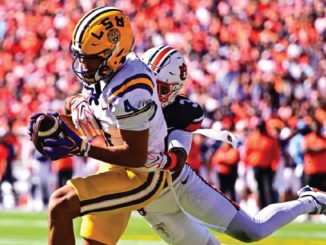 LSU Tigers player in purple and gold dives with the football as a defender in white chases behind on a crowded stadium field.
