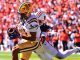 LSU Tigers player in purple and gold dives with the football as a defender in white chases behind on a crowded stadium field.