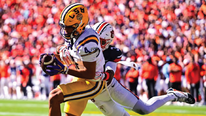 LSU Tigers player in purple and gold dives with the football as a defender in white chases behind on a crowded stadium field.