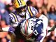 LSU player in a yellow helmet tackles Alabama player in navy uniform during a college football game.