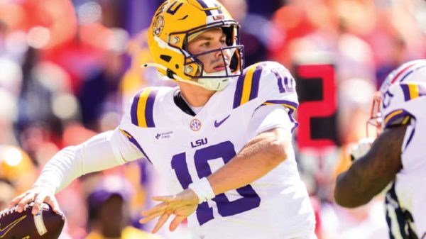 LSU quarterback in white jersey #10 and yellow helmet throwing a pass during a game.
