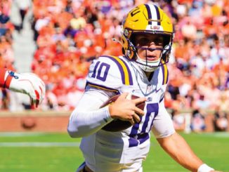 LSU quarterback wearing #10 in white and purple jersey runs with football on a green field during a game, crowd in orange in the stands behind him.