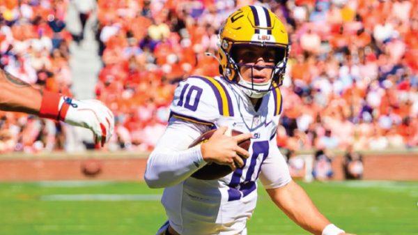 LSU quarterback wearing #10 in white and purple jersey runs with football on a green field during a game, crowd in orange in the stands behind him.