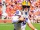 LSU quarterback wearing #10 in white and purple jersey runs with football on a green field during a game, crowd in orange in the stands behind him.