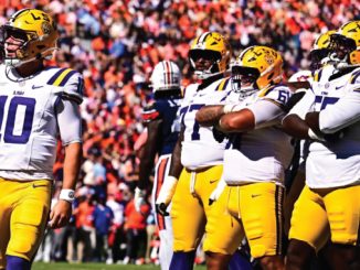 LSU football players in white jerseys and gold pants on a sunny sideline, quarterback number 10 in foreground amid teammates with gold helmets.