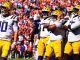 LSU football players in white jerseys and gold pants on a sunny sideline, quarterback number 10 in foreground amid teammates with gold helmets.