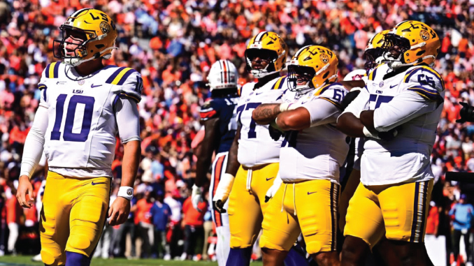 LSU football players in white jerseys and gold pants on a sunny sideline, quarterback number 10 in foreground amid teammates with gold helmets.