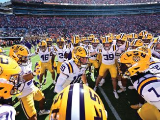 LSU football players form a huddle on the field in white uniforms and yellow helmets, stadium packed with fans behind them.
