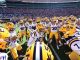 LSU football players form a huddle on the field in white uniforms and yellow helmets, stadium packed with fans behind them.