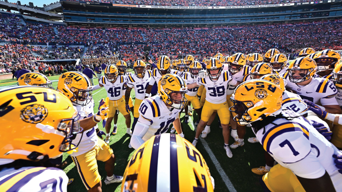 LSU football players form a huddle on the field in white uniforms and yellow helmets, stadium packed with fans behind them.