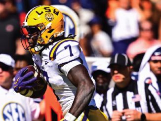 LSU football running back in white and purple uniform carries the ball during a game, yellow helmet with LSU logo and SEC crowd in the background.