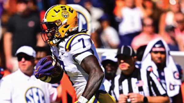 LSU football running back in white and purple uniform carries the ball during a game, yellow helmet with LSU logo and SEC crowd in the background.