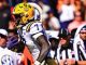 LSU football running back in white and purple uniform carries the ball during a game, yellow helmet with LSU logo and SEC crowd in the background.