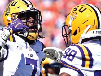 Two LSU football players in white jerseys and yellow helmets face off on the field during a game.