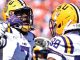 Two LSU football players in white jerseys and yellow helmets face off on the field during a game.
