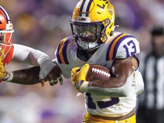 LSU running back (#13) cradles the football as a Clemson defender closes in during a college football play.