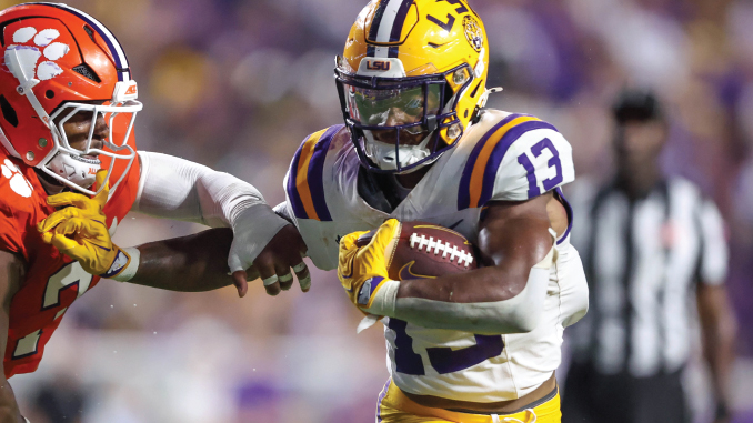 LSU running back (#13) cradles the football as a Clemson defender closes in during a college football play.