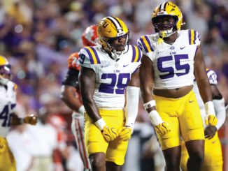 LSU Tigers football players in white and gold uniforms stand on the field during a game with a blurred crowd behind them.