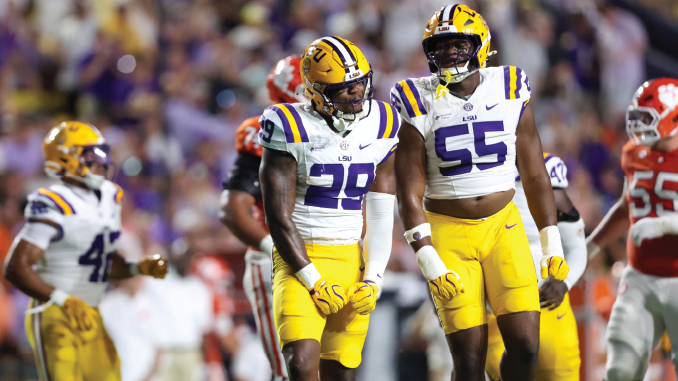 LSU Tigers football players in white and gold uniforms stand on the field during a game with a blurred crowd behind them.