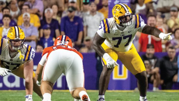 LSU offensive linemen (#77 and #57) in yellow helmets block a Clemson defender during a college football game as a crowd watches.