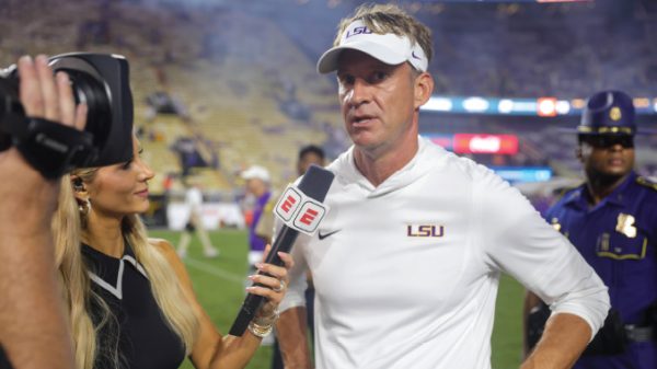 LSU coach in white polo and visor being interviewed on a football field by a reporter holding an ESPN microphone; cameras in view.
