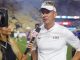 LSU coach in white polo and visor being interviewed on a football field by a reporter holding an ESPN microphone; cameras in view.