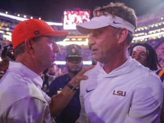 Two LSU coaches in white gear speak closely to each other in a crowded stadium tunnel after a game, one wearing an orange cap and the other a white visor.
