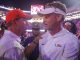 Two LSU coaches in white gear speak closely to each other in a crowded stadium tunnel after a game, one wearing an orange cap and the other a white visor.