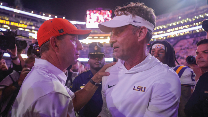 Two LSU coaches in white gear speak closely to each other in a crowded stadium tunnel after a game, one wearing an orange cap and the other a white visor.