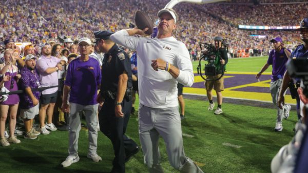 LSU quarterback in white throws a football on the sideline during a celebration on a packed stadium field, surrounded by fans, coaches, and photographers.