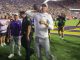 LSU quarterback in white throws a football on the sideline during a celebration on a packed stadium field, surrounded by fans, coaches, and photographers.