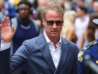 Man in a blue pinstripe suit and sunglasses raises his right hand as he walks past a crowd and a security officer nearby.