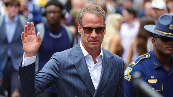 Man in a blue pinstripe suit and sunglasses raises his right hand as he walks past a crowd and a security officer nearby.