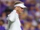 Coach or athlete wearing a white polo and visor on a sports field, cupping his ear toward the crowd.